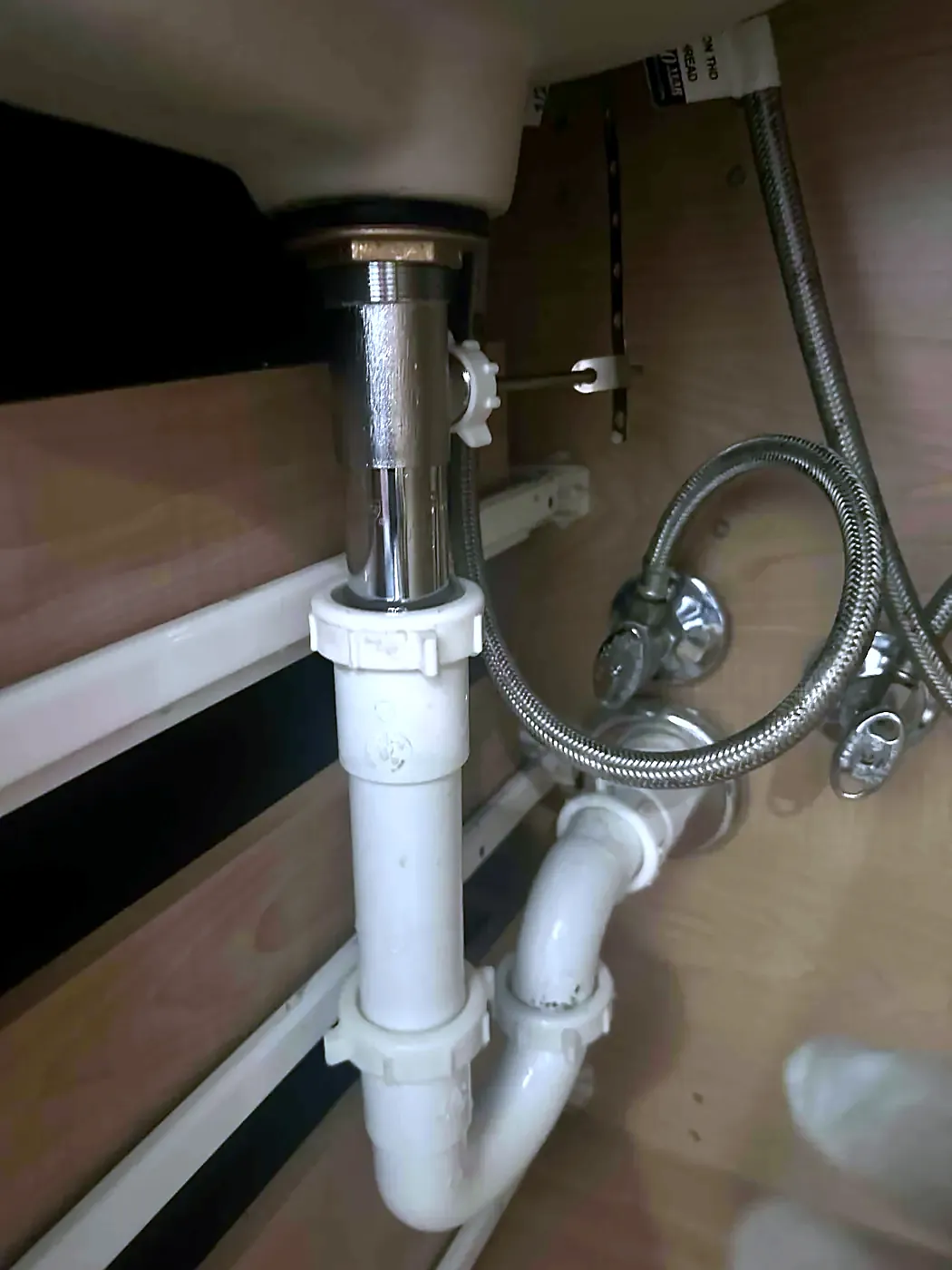 Under-sink view of white PVC P-trap, chrome tailpiece and braided supply lines inside a wood cabinet