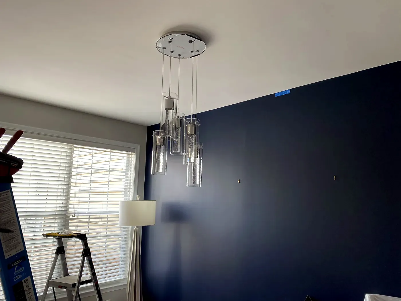 Cluster of glass cylinder pendants hanging from one canopy beside a dark navy wall, stepladder at the window