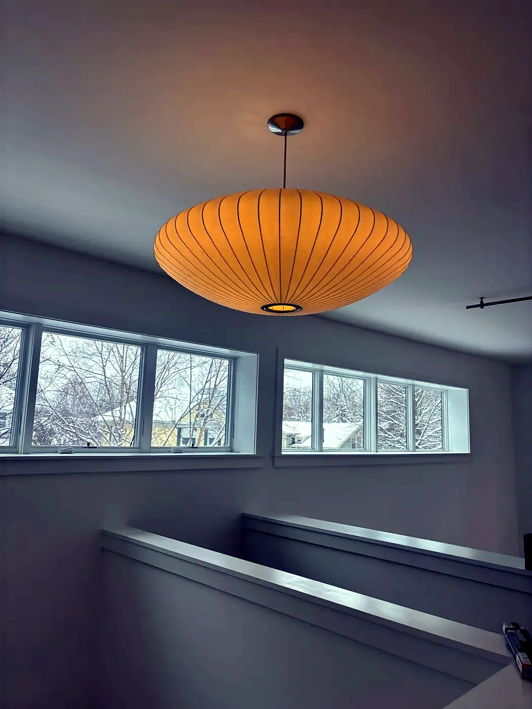 Glowing ribbed saucer-shaped pendant light hanging in a loft with a row of windows and a snowy view outside