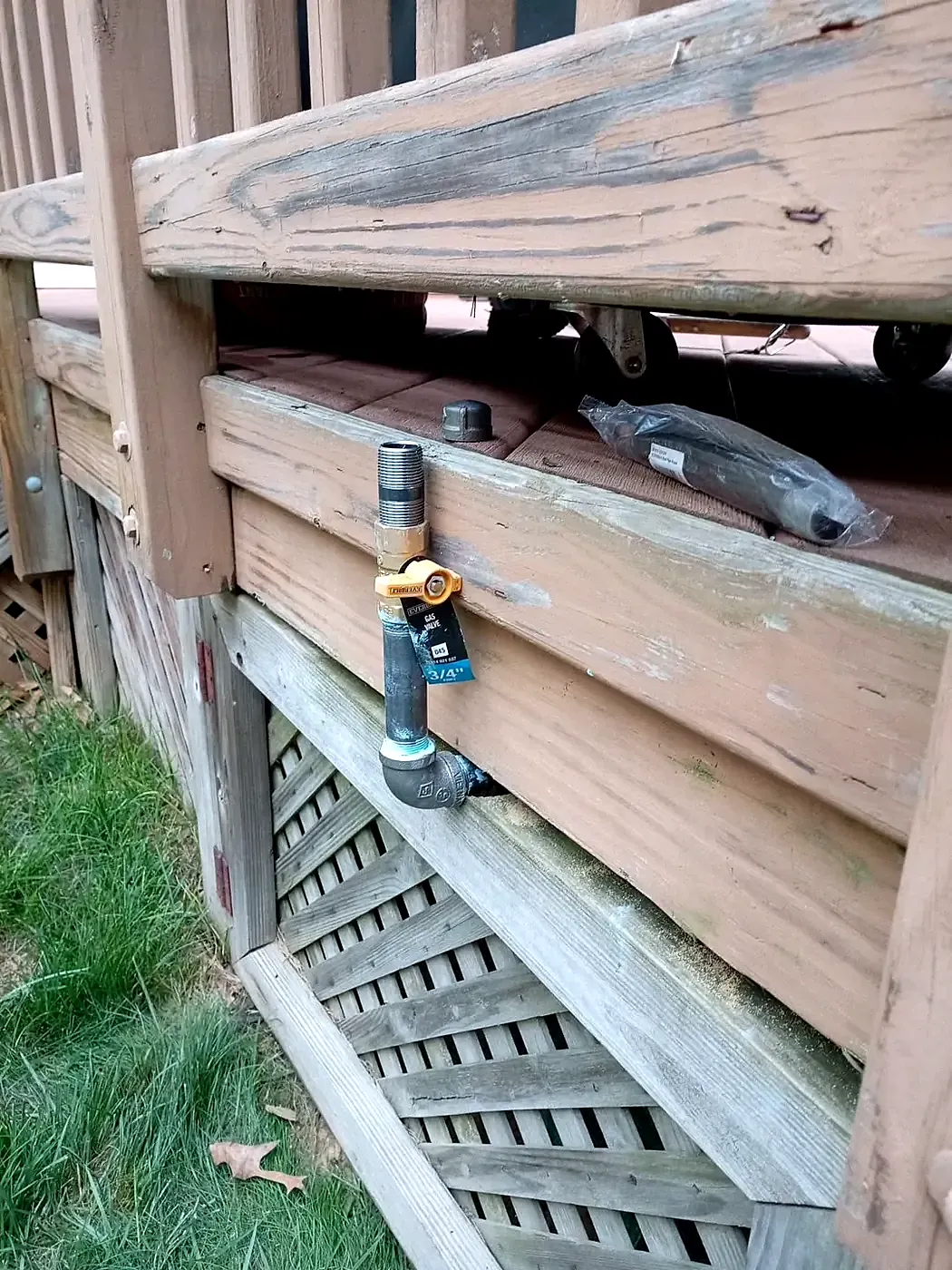 Brass hose bib mounted on a weathered wood deck rim above lattice skirting and grass