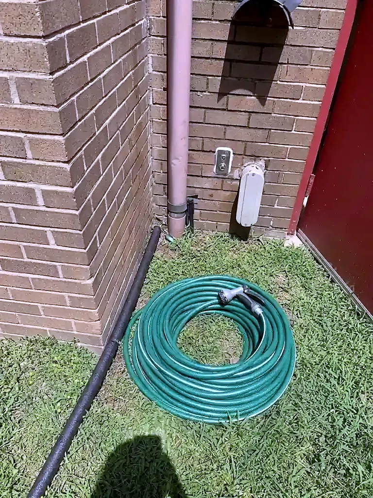 Hose bib on a brick wall beside a downspout with a green garden hose coiled on the lawn