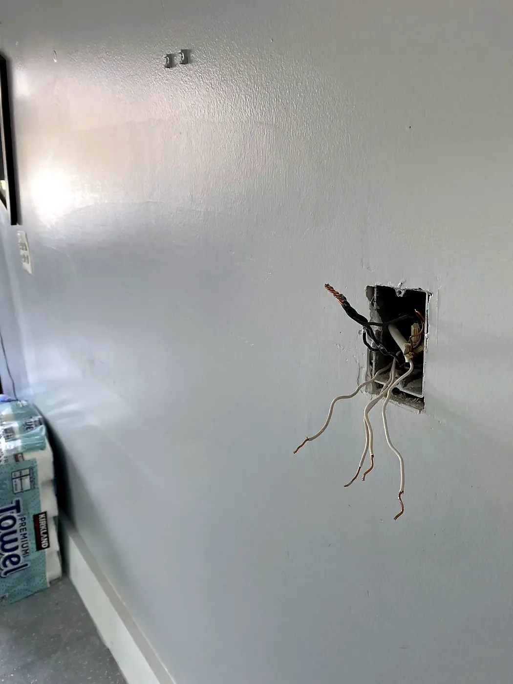 Bare wires hanging from a small rectangular opening in a gray painted wall