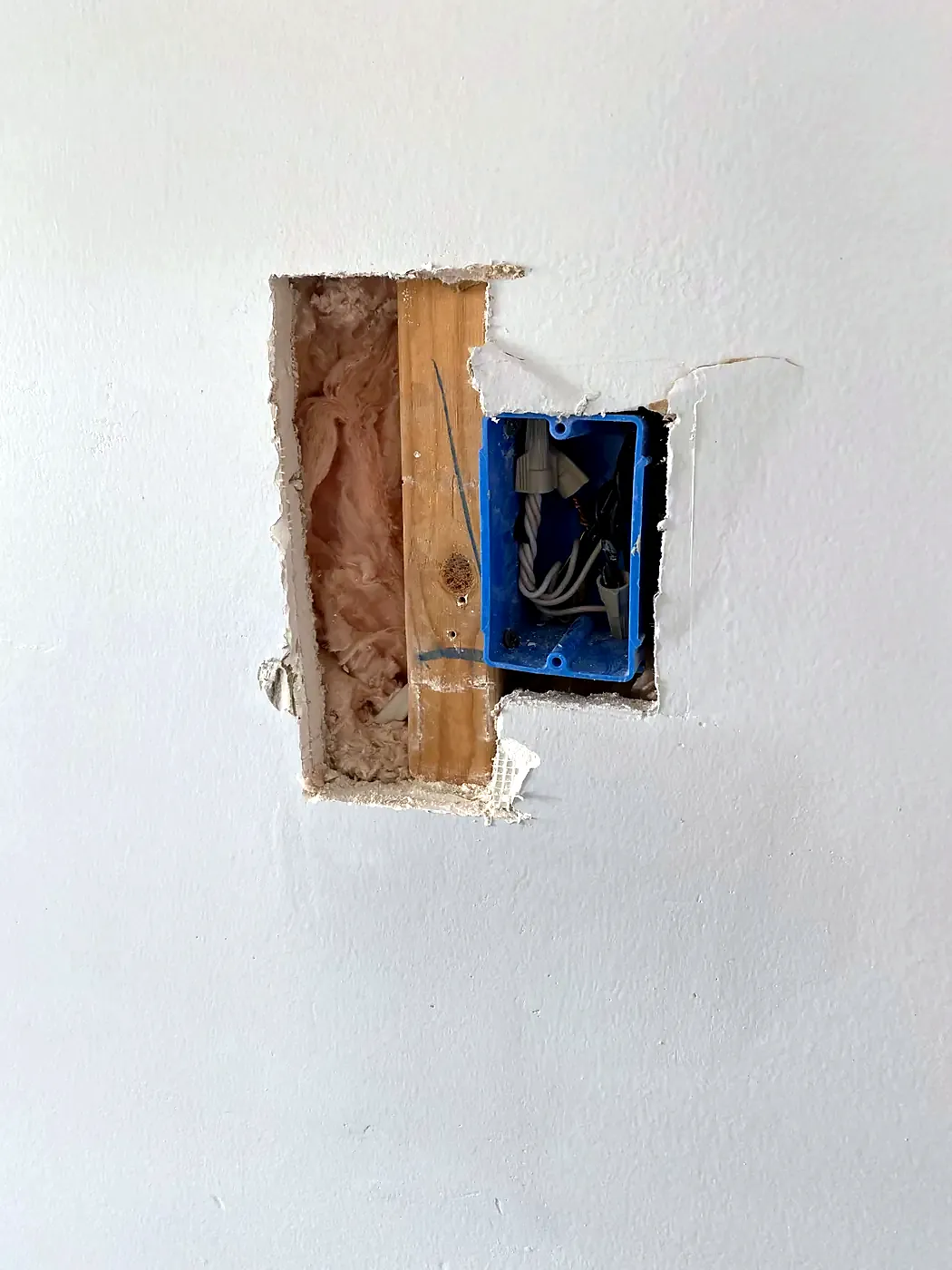Rectangular drywall cutout showing a blue electrical box, wires, a wood stud and insulation