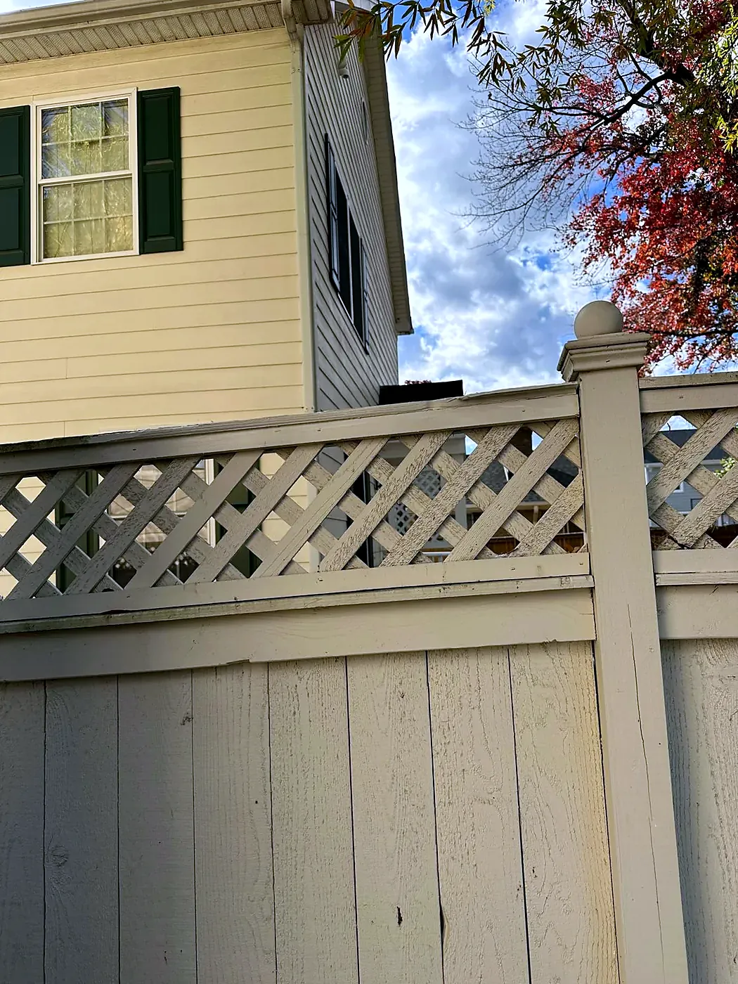 Cream painted lattice-top fence beside a yellow-sided house with dark green shutters under a cloudy sky