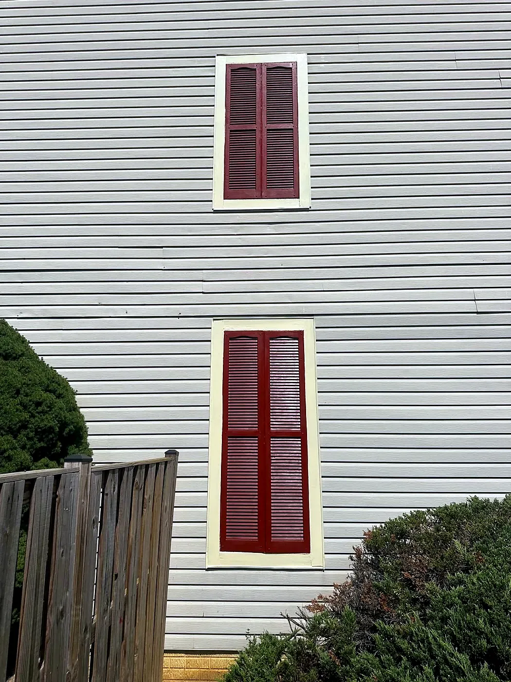 Two windows with red shutters on a freshly painted gray-sided wall above green shrubs