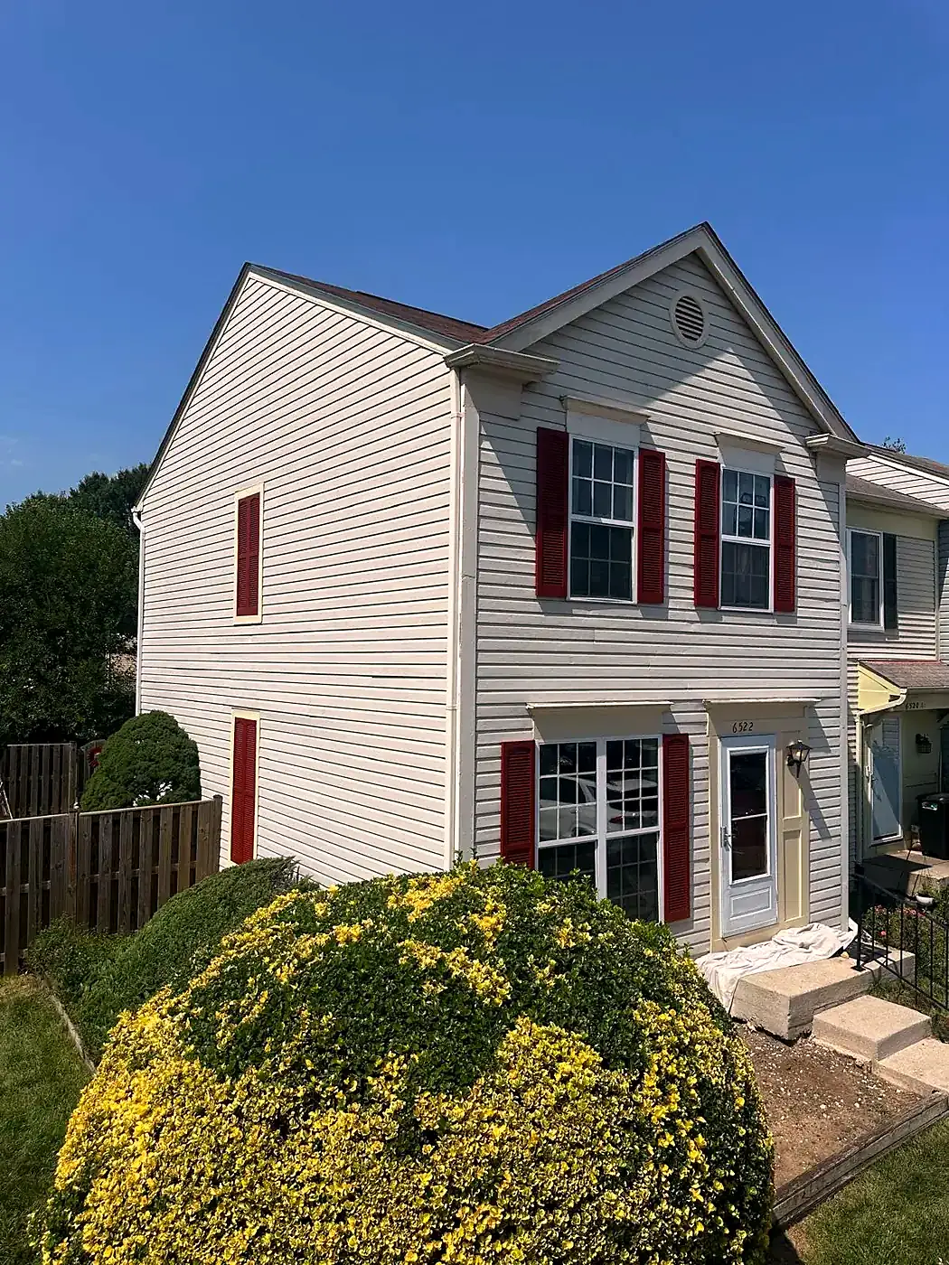 Two-story townhouse with gray siding, red shutters and white trim behind a round yellow shrub under blue sky