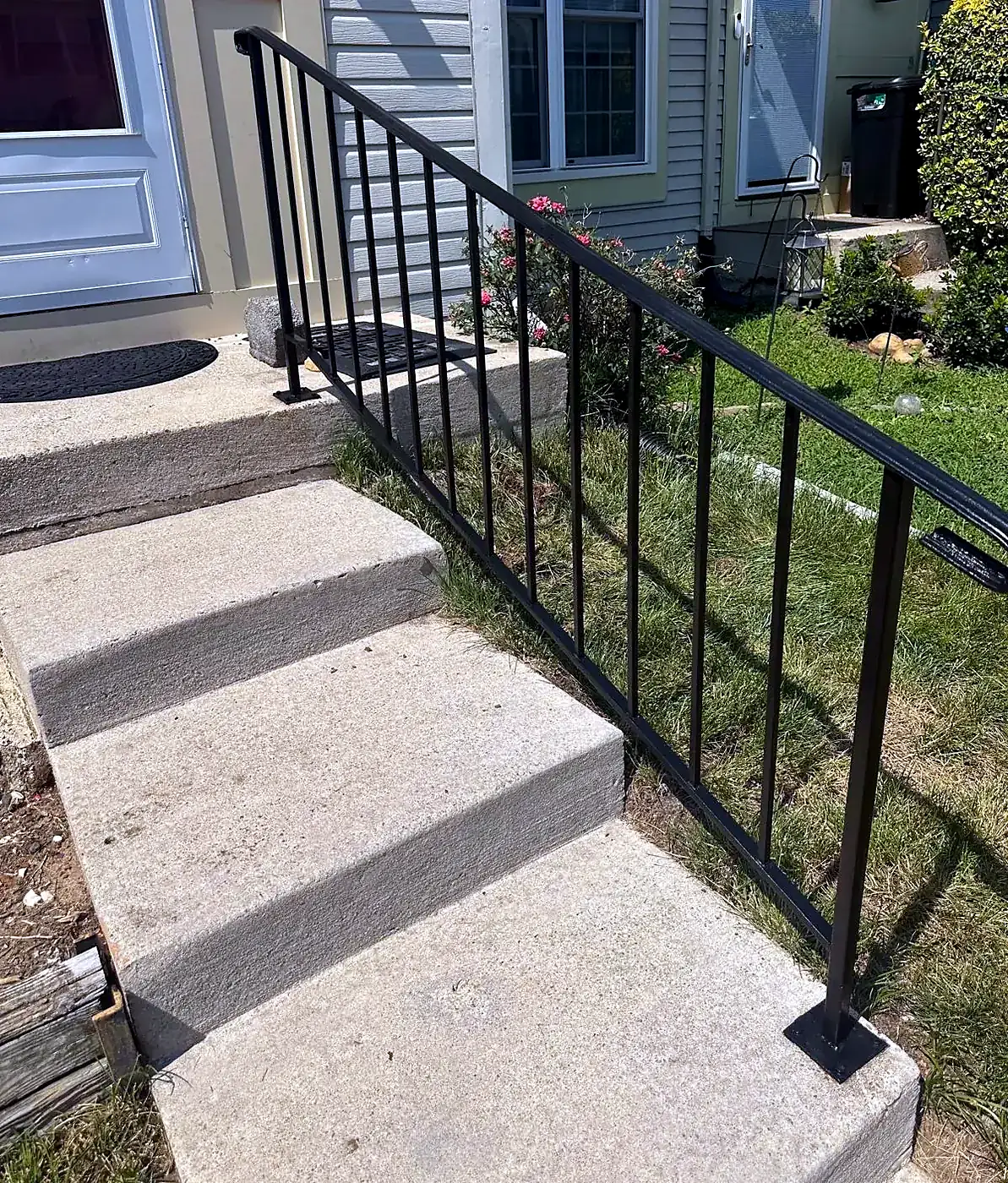 Black painted metal handrail on concrete front steps beside a house with tan siding and a white door