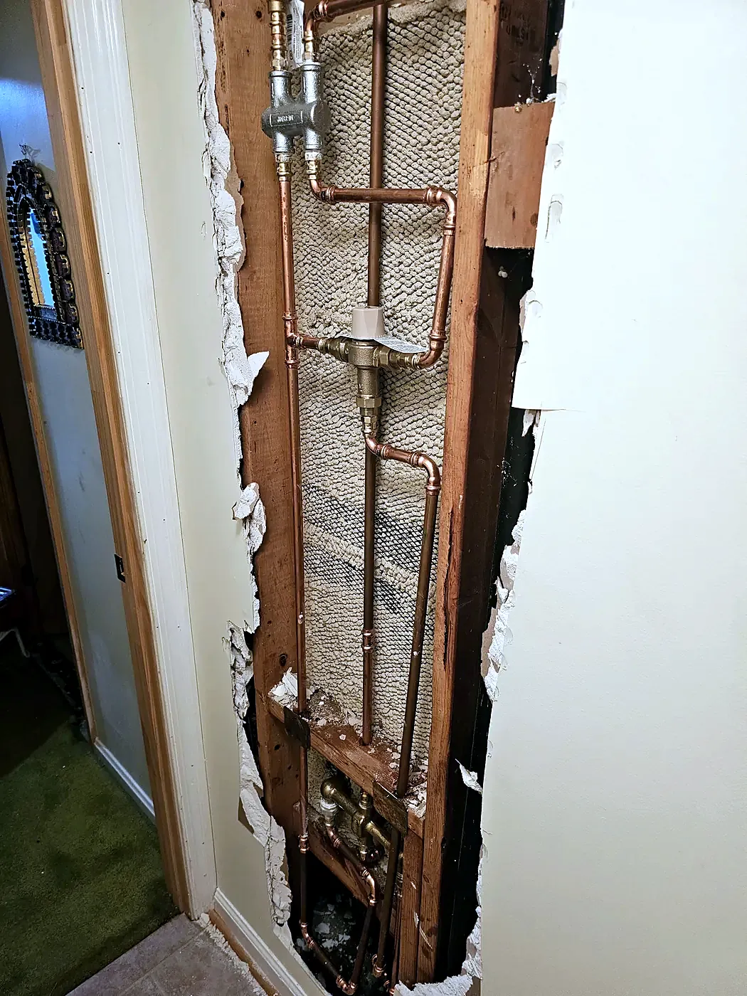 Narrow vertical opening in a hallway wall revealing copper pipes and valves between wood studs