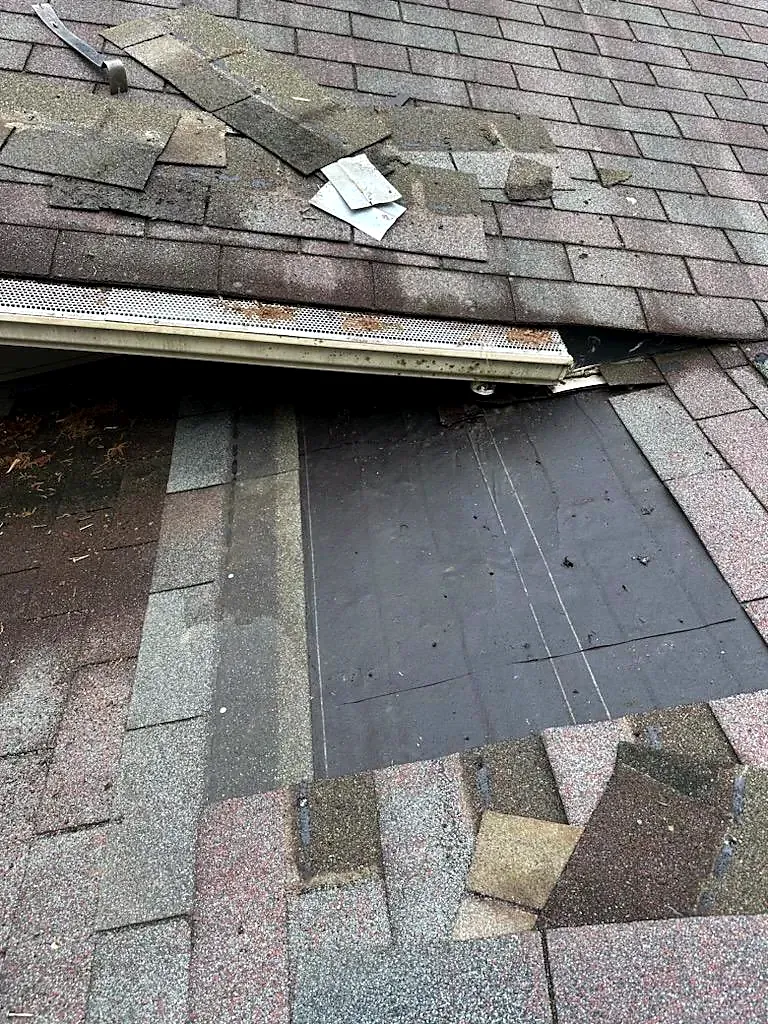 Torn and lifted asphalt shingles near a gutter with underlayment exposed and loose pieces