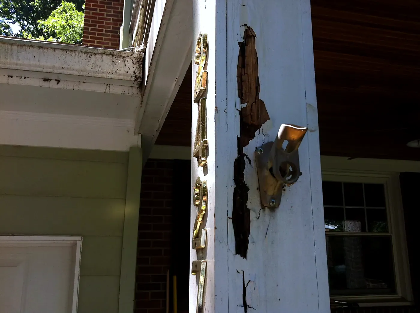 White porch column with splitting rotted wood and brass hinge hardware beside a brick wall and front door