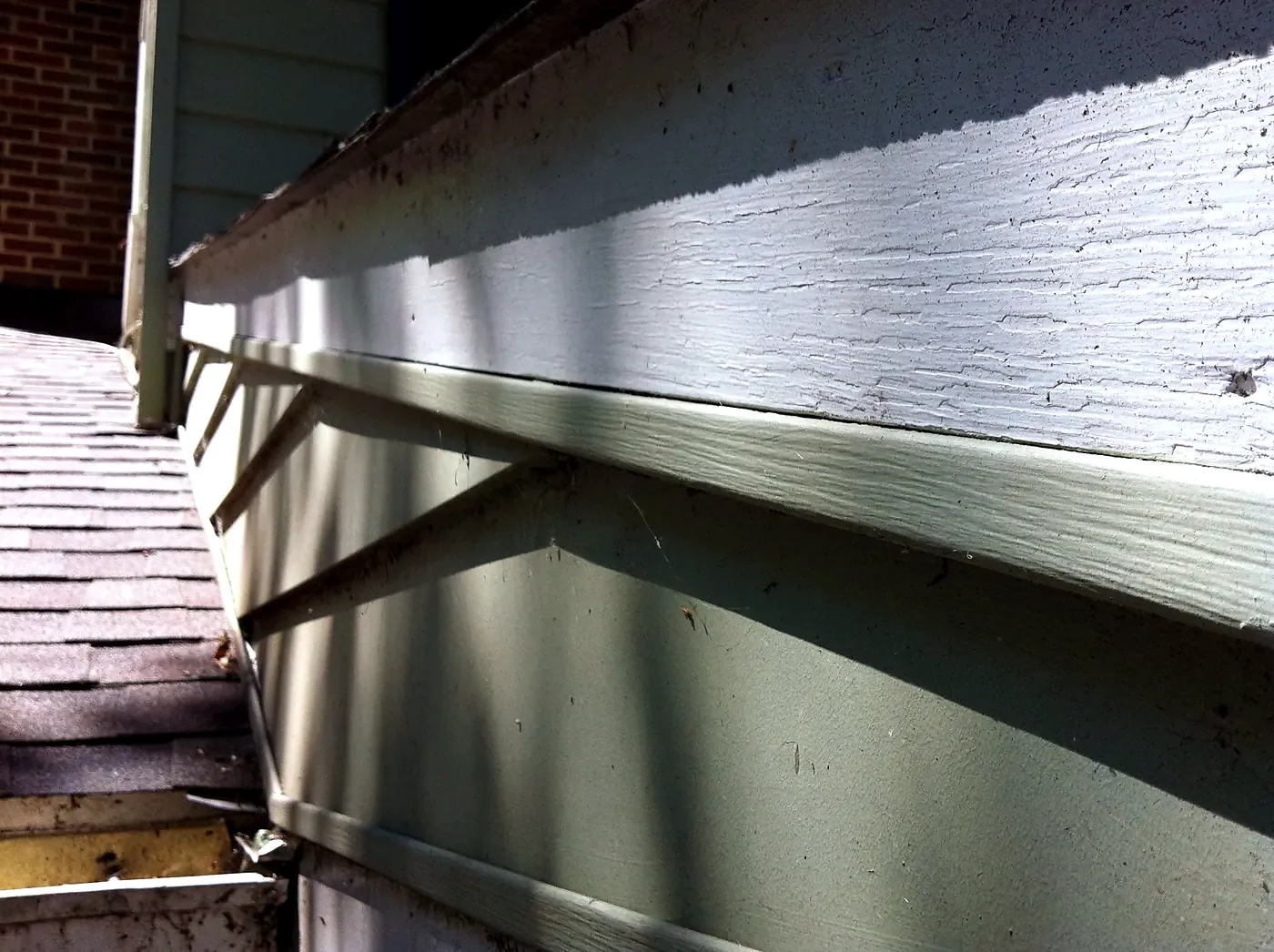 Weathered white fascia board with peeling paint along a shingle roof edge above gray siding