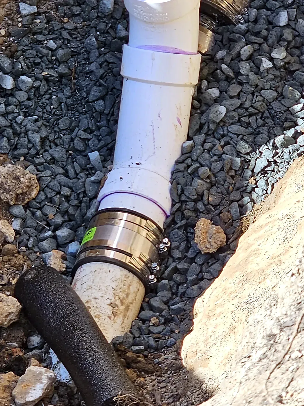 White PVC pipe joined to a black pipe with a metal-banded coupling, set in gray gravel