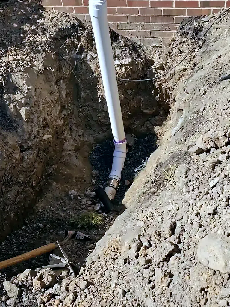 Deep dirt trench against a brick house wall with a white PVC pipe rising from gravel at the bottom