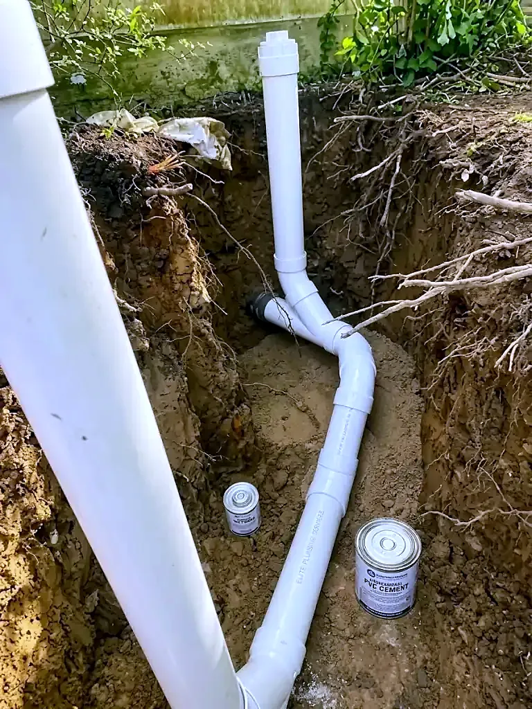 White PVC sewer pipe with a vertical cleanout riser in a dirt trench, two cans of pipe cement beside it