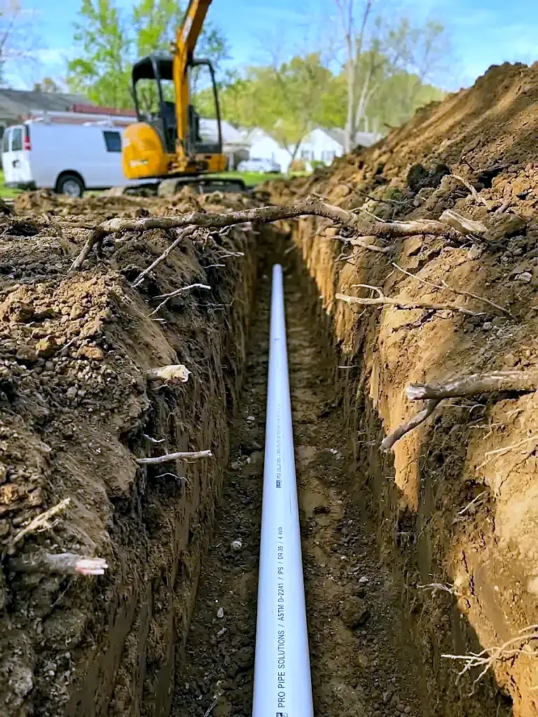 Narrow deep trench with a white PVC sewer pipe at the bottom, yellow mini excavator at the far end