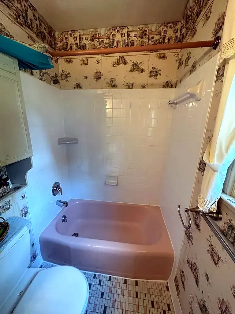 Old pink bathtub with white tile walls and floral wallpaper in a small bathroom