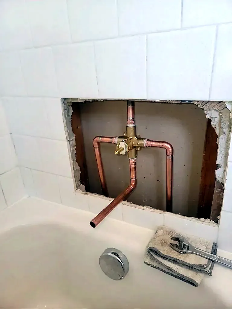 Opening in a white tile wall showing a new brass valve body and copper pipes above a tub