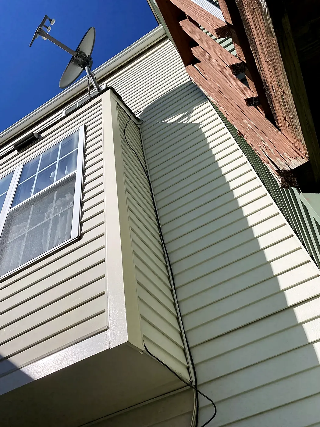 Upward view of cream vinyl siding under a deck with rotted ledger framing and a satellite dish, blue sky
