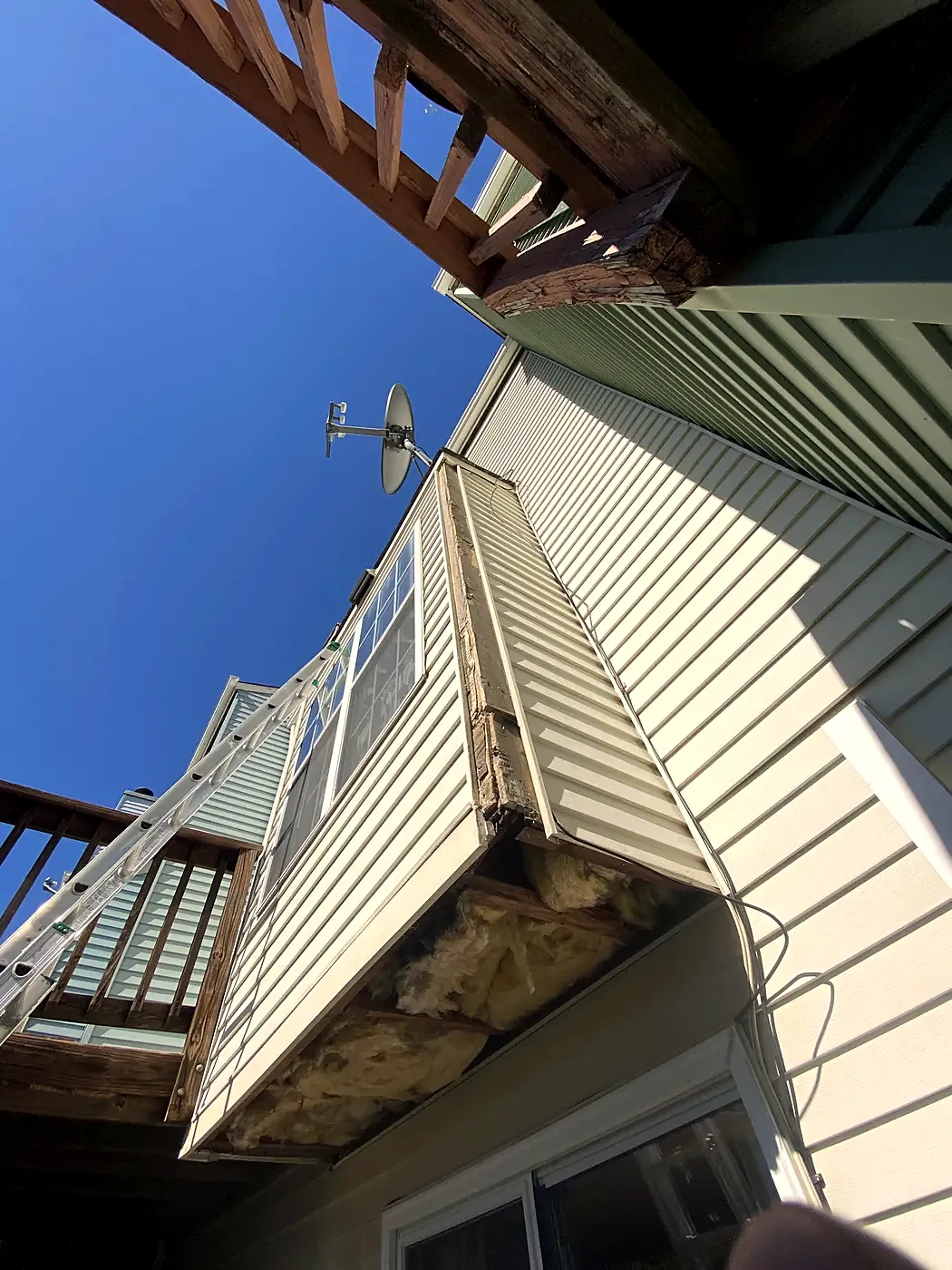 Vinyl siding damage under a wood deck with insulation exposed below a bay window, seen from below