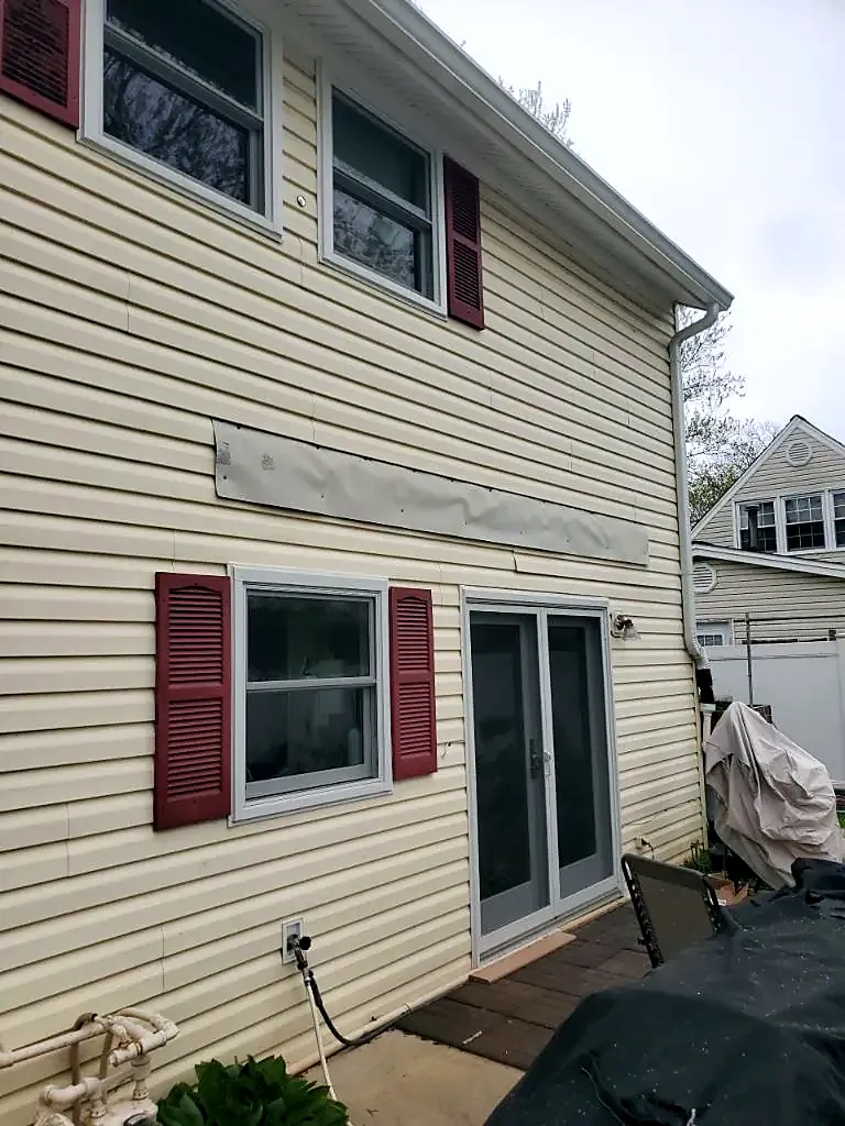 Rear of a cream-sided house with maroon shutters, a patched strip above the sliding door and a tarp on the deck