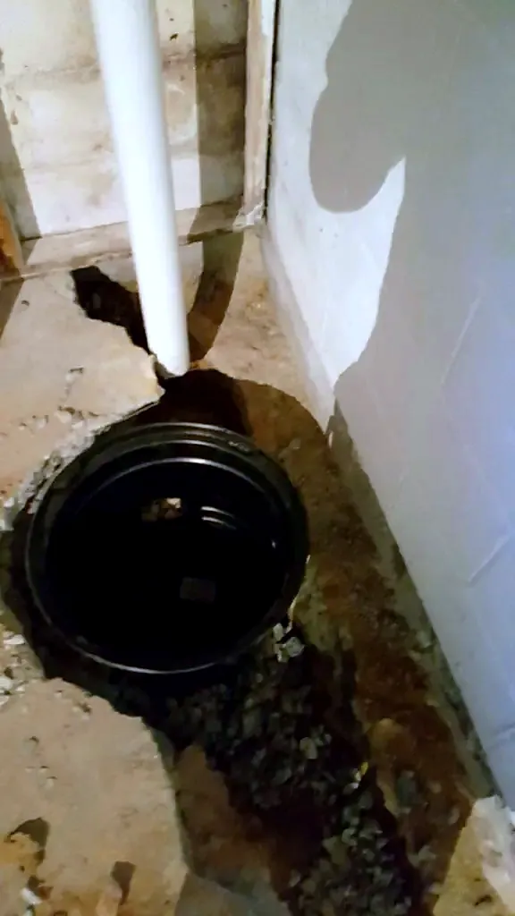 Open sump basin in a stained basement floor corner with a white discharge pipe rising along the block wall