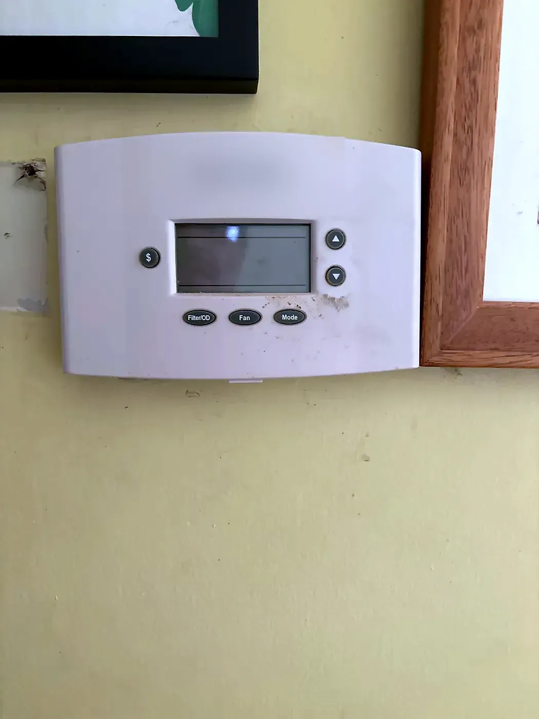 White programmable thermostat with small display on a pale yellow wall next to a wood picture frame