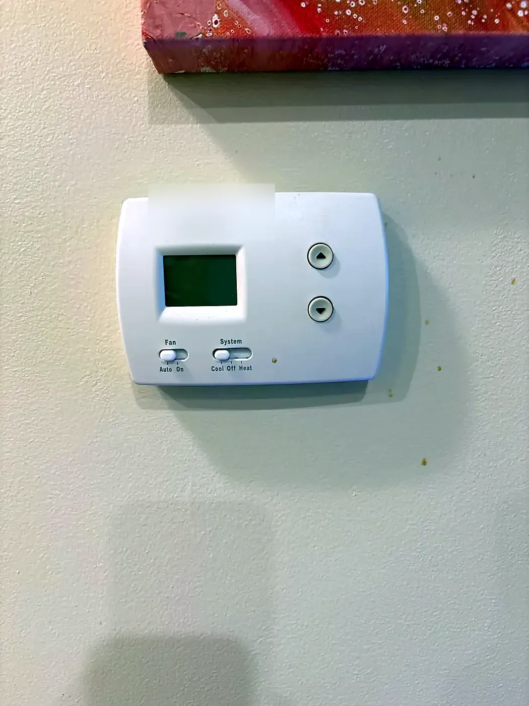 Simple white thermostat with small screen and slide switches on a beige wall below a pink canvas