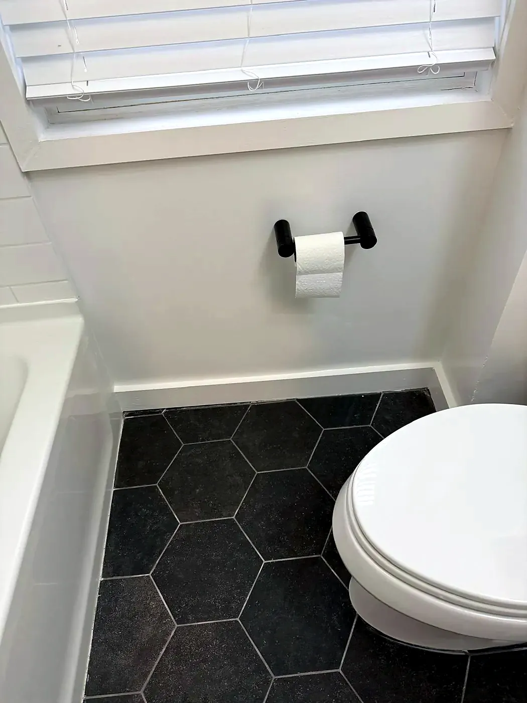 Repaired dark hexagon tile floor between a white bathtub and toilet, under a window
