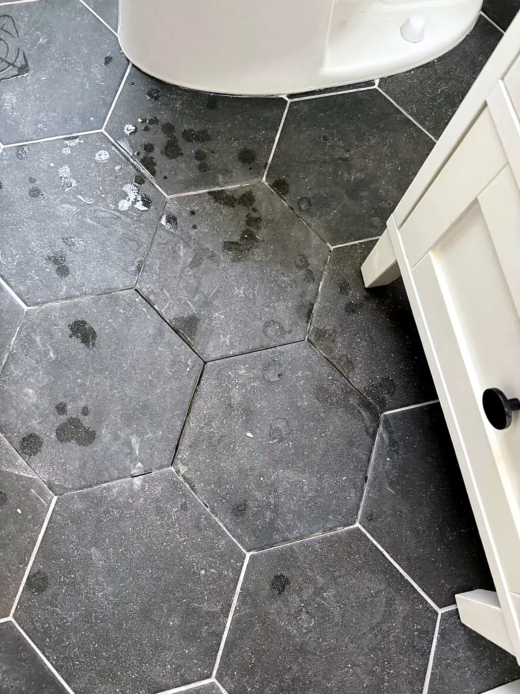 Dirty dark hexagon tile floor with stains and chipped grout beside a white cabinet