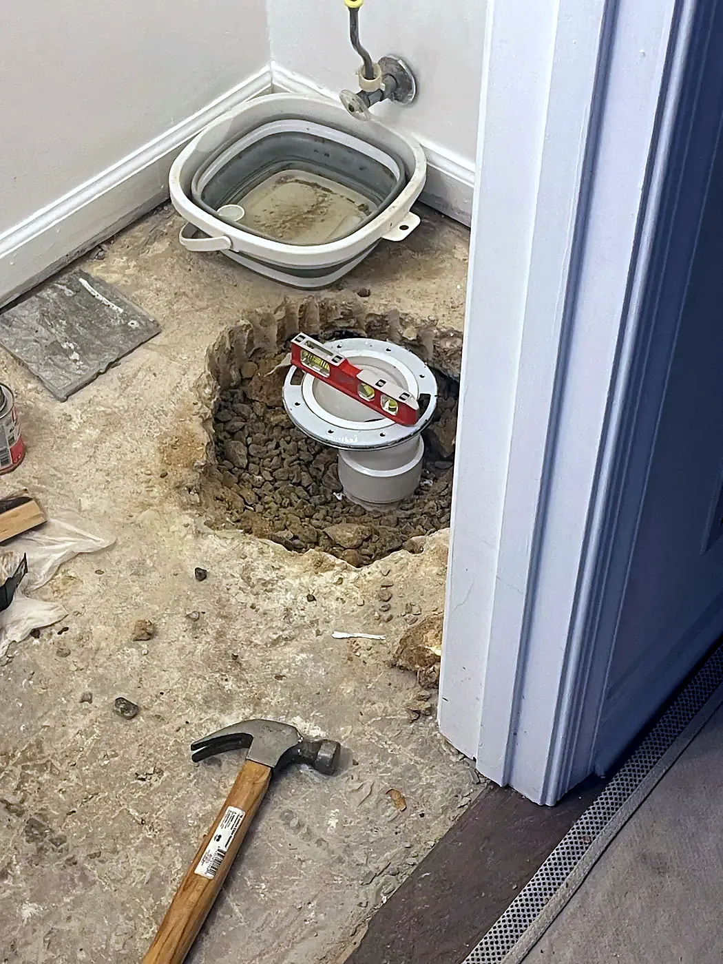 Bathroom floor removed around a toilet flange with gravel, a hammer and a bucket