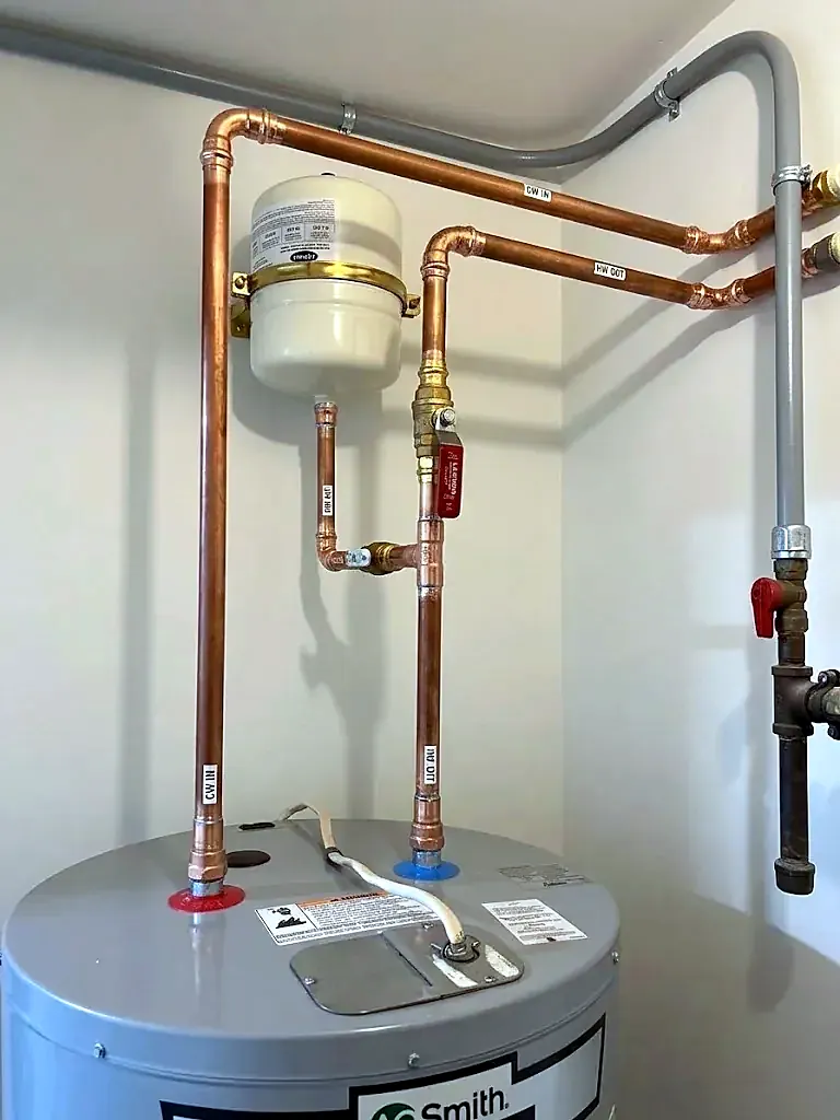 Top of a gray water heater with new copper piping, red ball valves and a white expansion tank on the wall
