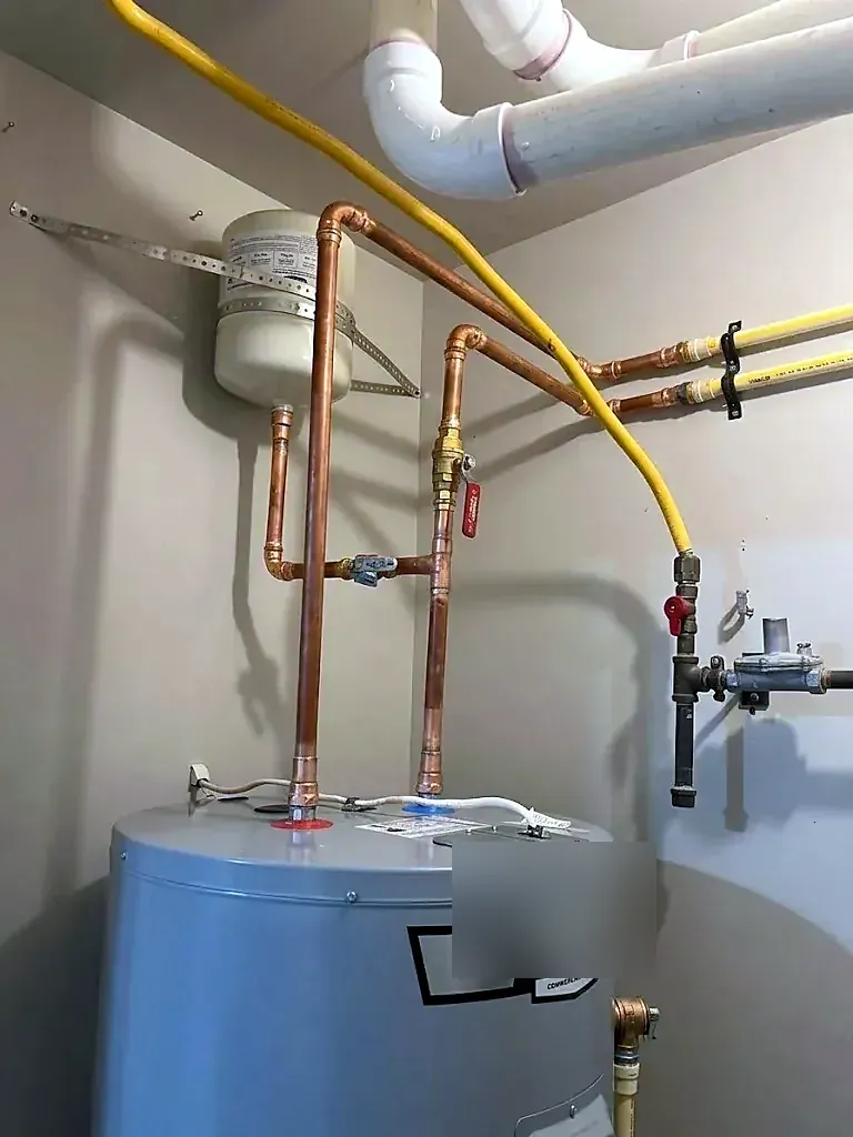 Gray water heater with copper lines, an overhead expansion tank and a yellow gas connector beside PVC piping
