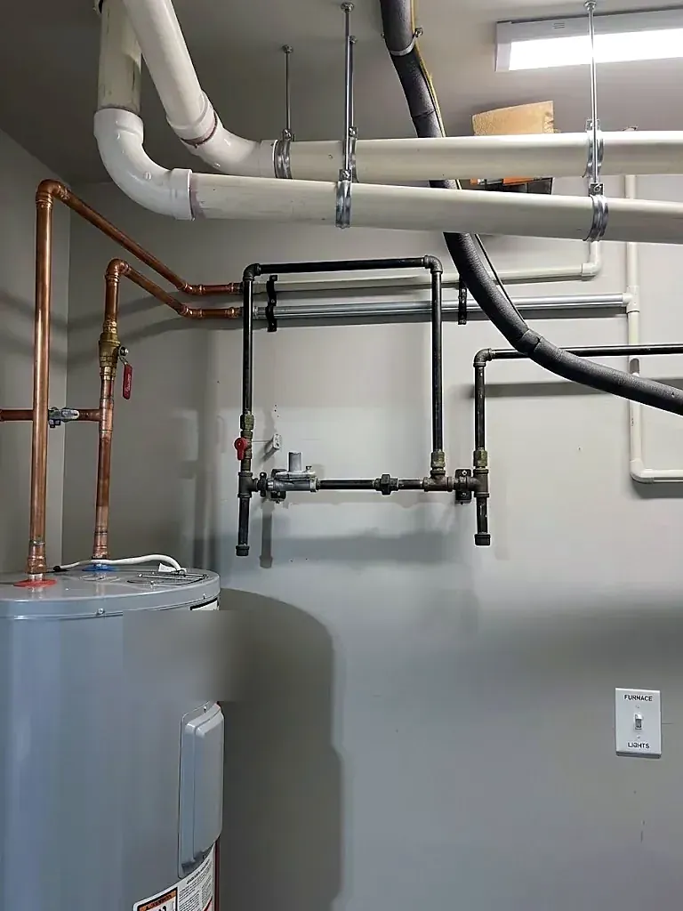 Copper water lines running from a gray water heater across a wall with black iron gas pipe and PVC drains above