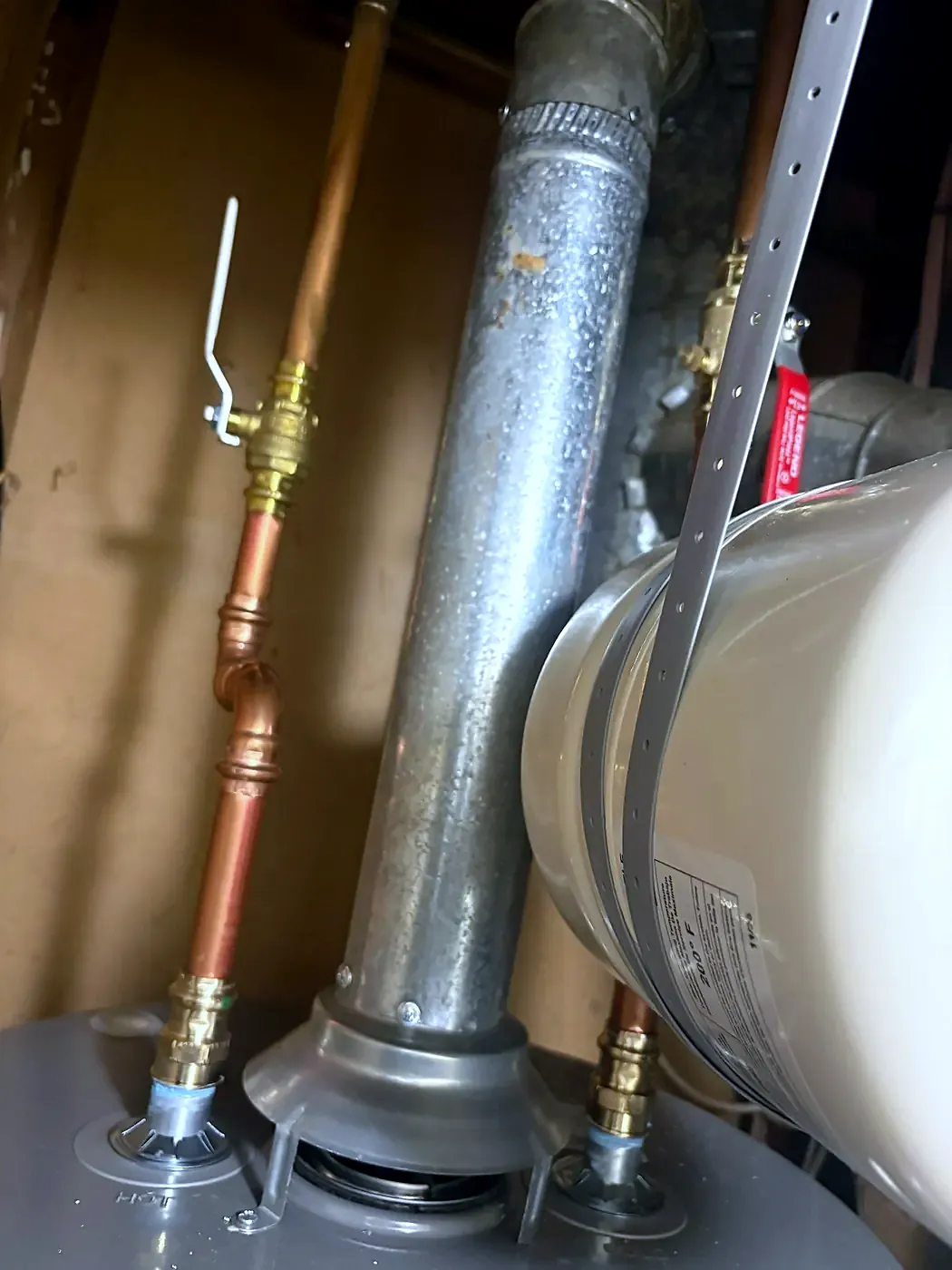 Top of a water heater with a metal flue, copper pipes and a gas valve against a wood-framed wall