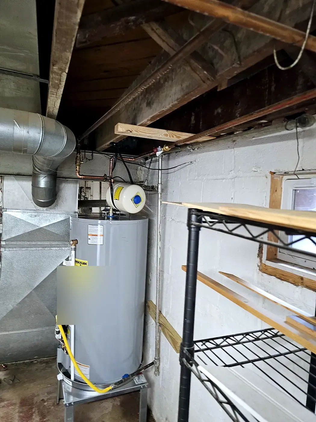 Gray tank water heater with yellow gas line beside a wire shelf in a basement with white block walls