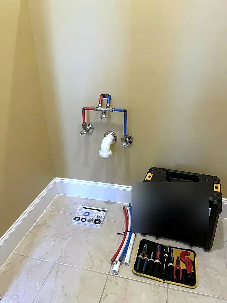 Red and blue PEX stubs and a drain roughed in at a laundry wall, toolbox on the tile floor