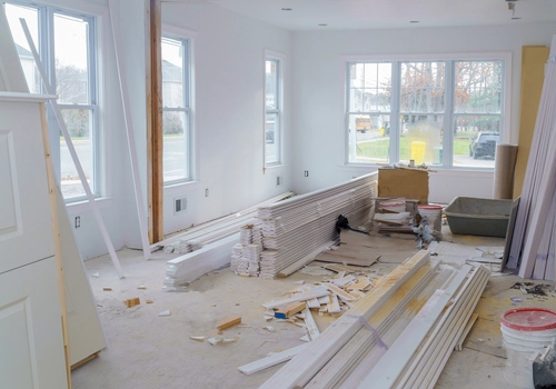 unfinished interior with stacks of white planks leaning against a wall. These planks could be drywall or trim boards. The room lacks any wall coverings or paint, revealing the bare structure. Large windows allow natural light to flood the space. The floor is strewn with debris, including wood pieces and sawdust. An exposed ladder on the left side suggests ongoing work, and electrical outlets without faceplates are visible.