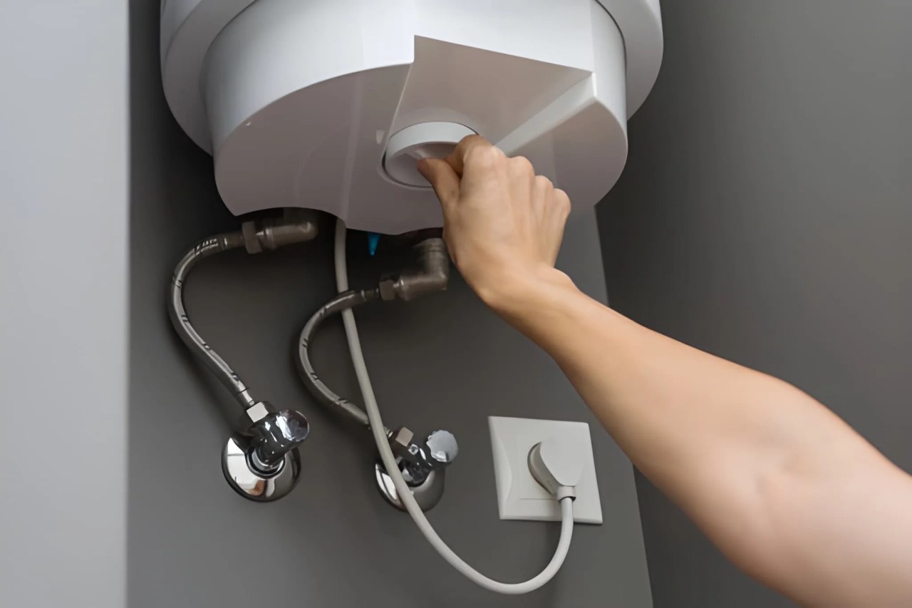 homeowner adjusting the temperature control knob on an electric water heater mounted under a sink.