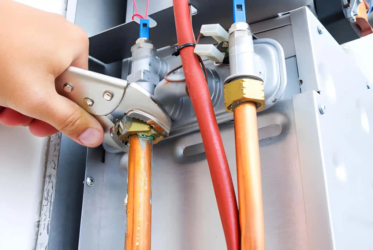 hand using a wrench to tighten a copper pipe connection inside a heating system, with red and orange pipes visible. furnace installation near me in ashburn va