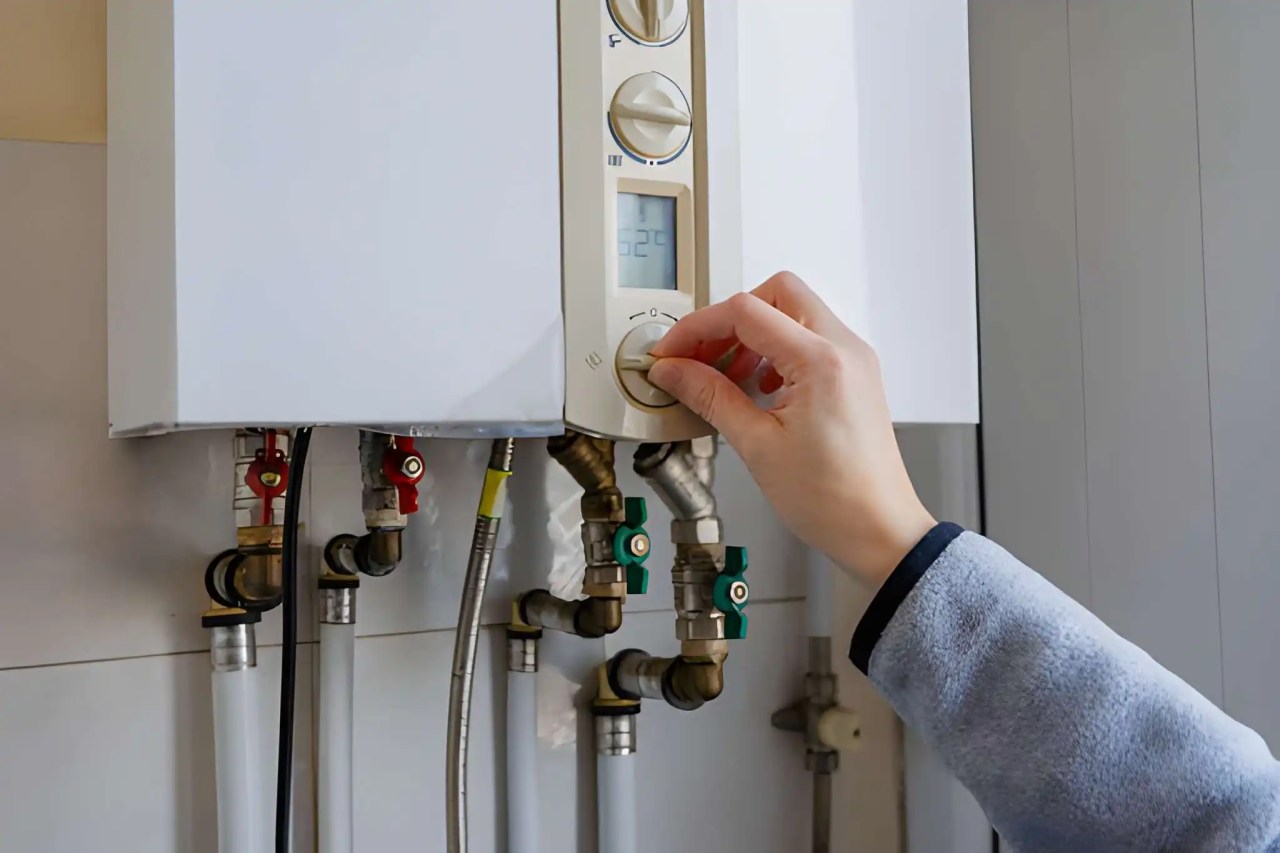 hand adjusting the control knob on a wall-mounted furnace or boiler unit, with visible pipes and shutoff valves underneath. furnace installation near me in fairfax station va