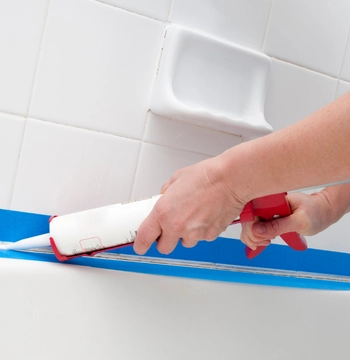 bathroom caulking repair in mclean va showing a professional applying a fresh bead of white silicone sealant to a bathtub rim using blue painter's tape for a clean, precision finish.