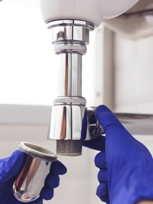 a detailed view of a professional bathroom sink installation focusing on a plumber in blue gloves carefully connecting a chrome bottle trap to the basin's drainage assembly.