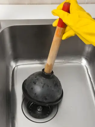 close-up of a clogged sink inspection in progress, showing a person in yellow gloves using a traditional rubber plunger to clear a blockage from a stainless steel kitchen sink.