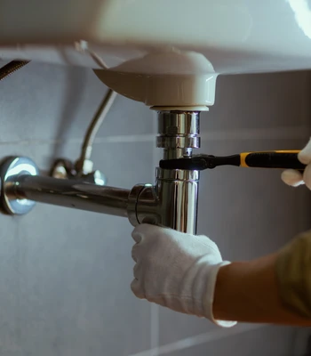 professional burst pipe repair in falls church, va, showing a plumber in white safety gloves using a specialized wrench to tighten a chrome p-trap drainage assembly beneath a bathroom sink.