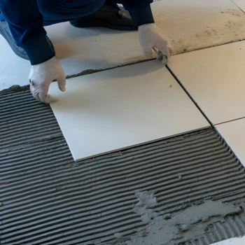 a skilled tile professional wearing protective white gloves carefully sets a large square white ceramic tile onto a freshly combed bed of grey thinset mortar during a residential floor tile repair burke va.