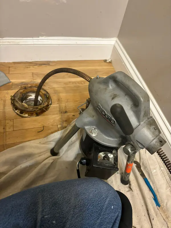 View looking down a protected hallway runner into a half bathroom with a vessel sink, where a portable electric drain cleaner machine is placed on a drop cloth next to a removed toilet area.