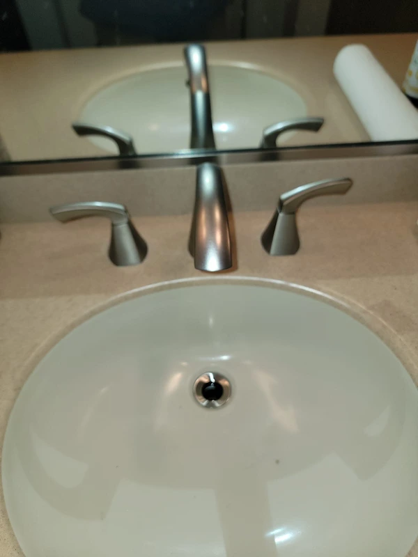 Straight-on view of a newly installed widespread brushed metal bathroom faucet reflected in a vanity mirror, highlighting clean pop-up drain integration and a spotless basin.