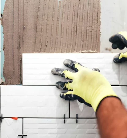 a professional tile repair contractor burke va wearing yellow work gloves to install a textured white subway tile onto a vertical wall backed with brown thinset mortar.